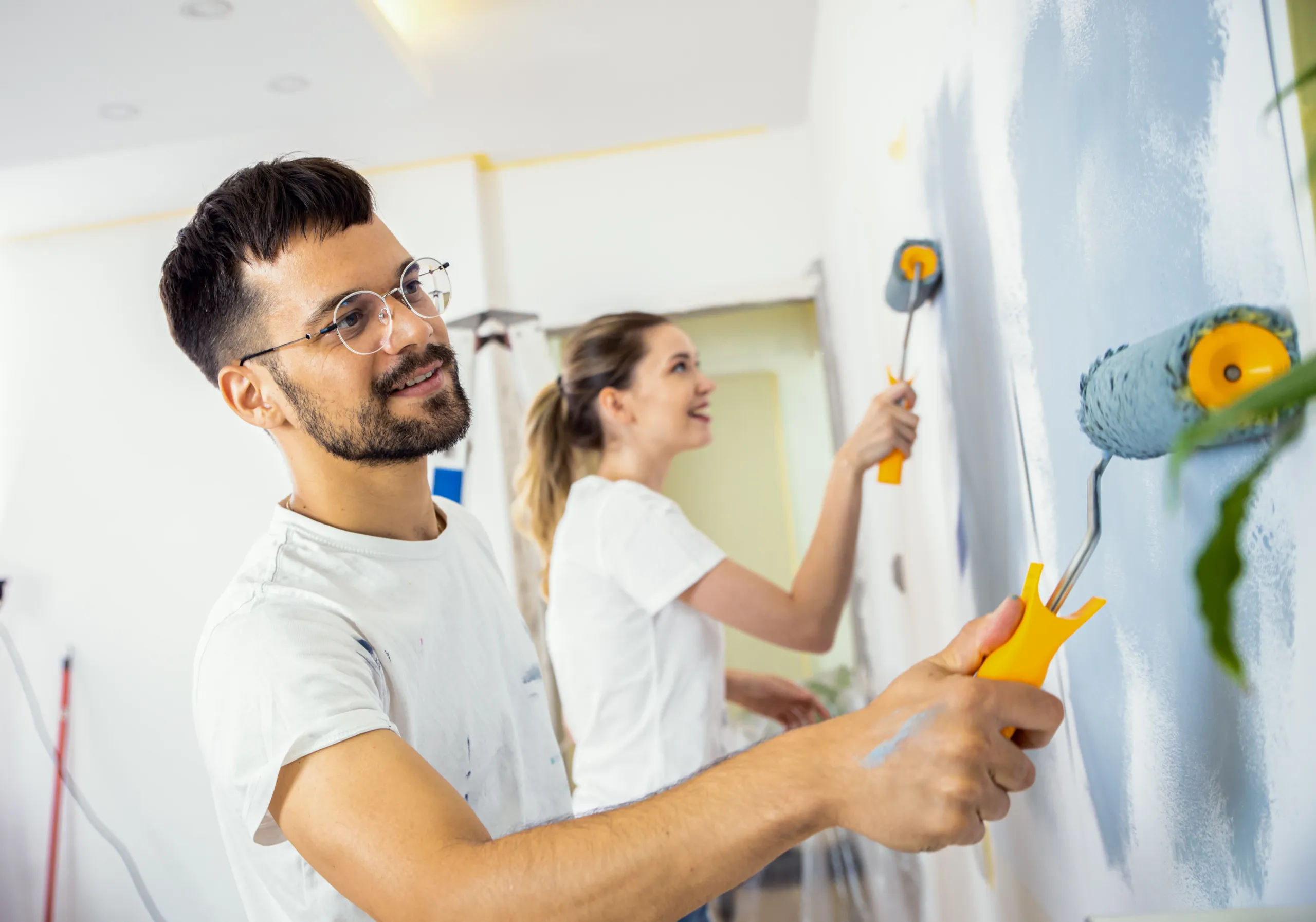 Two people painting a wall light blue with paint rollers in a bright room. Both are wearing white shirts and appear focused on the task.