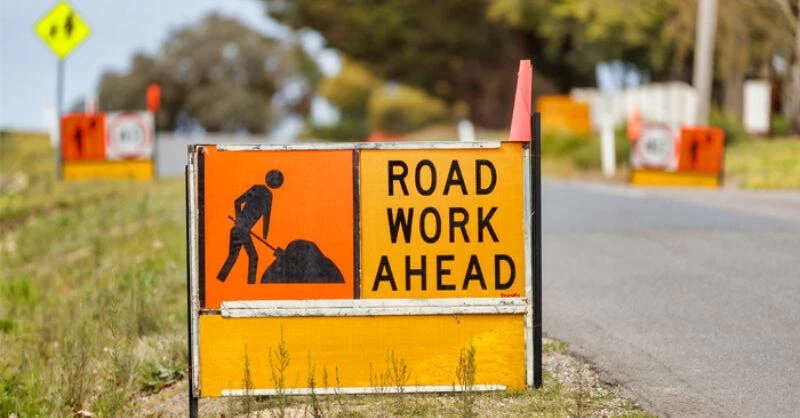 A yellow and orange sign by a roadside reads “ROAD WORK AHEAD” with an image of a person shovelling beside a pile of earth.