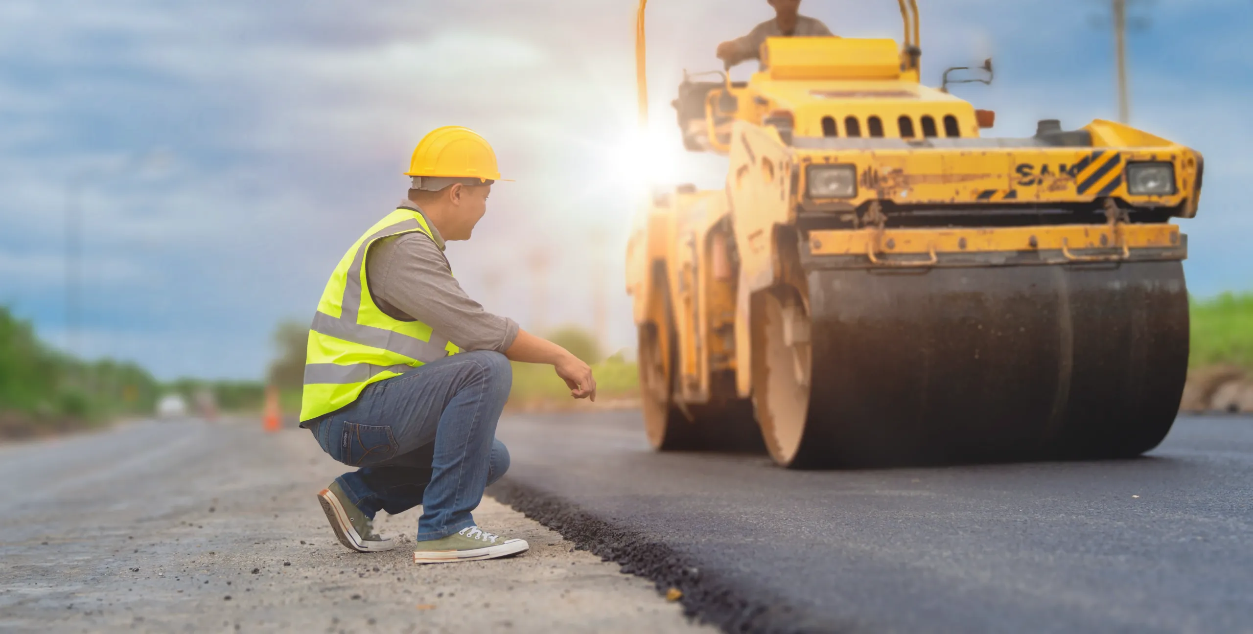 A construction worker in a safety vest and helmet inspects freshly laid tarmac as a road roller compacts the surface under a cloudy sky.